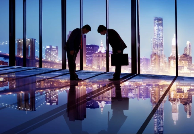 Two business professionals bowing in a glass-walled office overlooking an illuminated city skyline.
